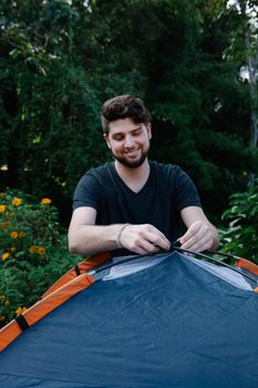 Cheerful bearded male traveler putting up tent during weekend trip against trees in daytime - Fotografer Sidoarjo