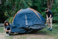 Couple of travelers setting up tent on meadow against trees and bushes during summer journey in daylight
