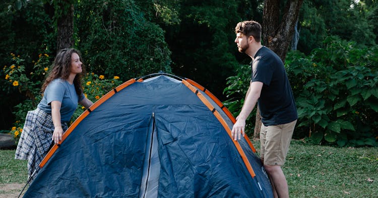 Positive Young Couple Setting Up Camp Tent In Forest