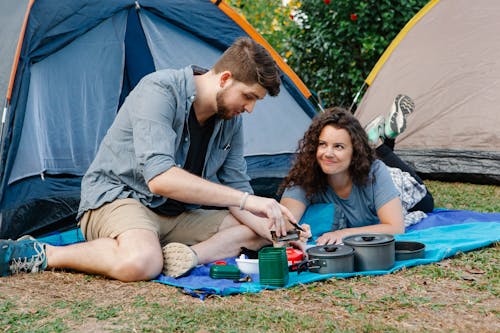 Positive young couple using portable camping gas stove while resting of plaid near tent during romantic trip in nature