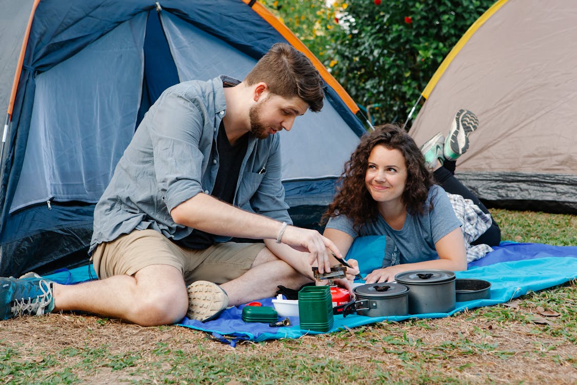 Positive young couple using portable camping gas stove while resting of plaid near tent during romantic trip in nature