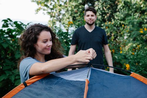 Free Happy young female traveler with long curly hair tying tent while standing near lush green trees during camping with boyfriend Stock Photo