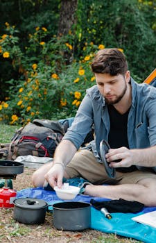A young man cooking outdoors at a campsite surrounded by vibrant flowers and camping gear.