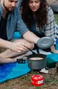 Crop young male and female campers in casual clothes cooking soup in saucepan on portable gas stove while resting on blanket after trekking in nature