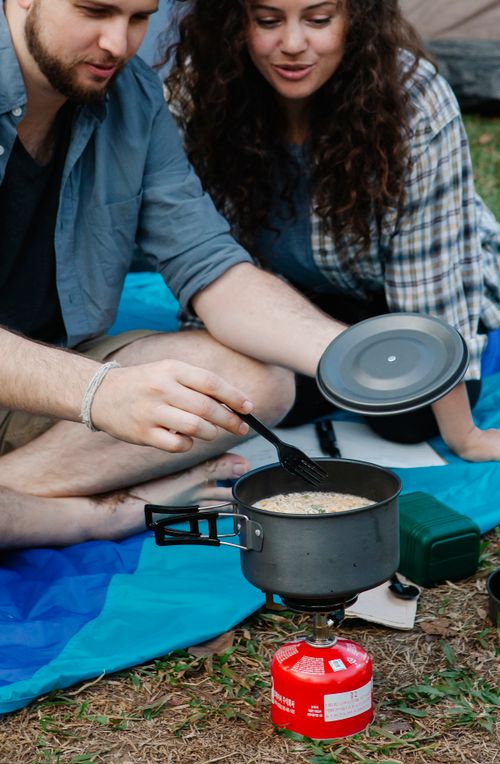 Free Crop young male and female campers in casual clothes cooking soup in saucepan on portable gas stove while resting on blanket after trekking in nature Stock Photo