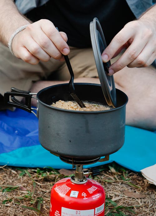 Crop unrecognizable male traveler in casual clothes stirring hot dish in saucepan on gas stove while preparing dinner during camping in nature