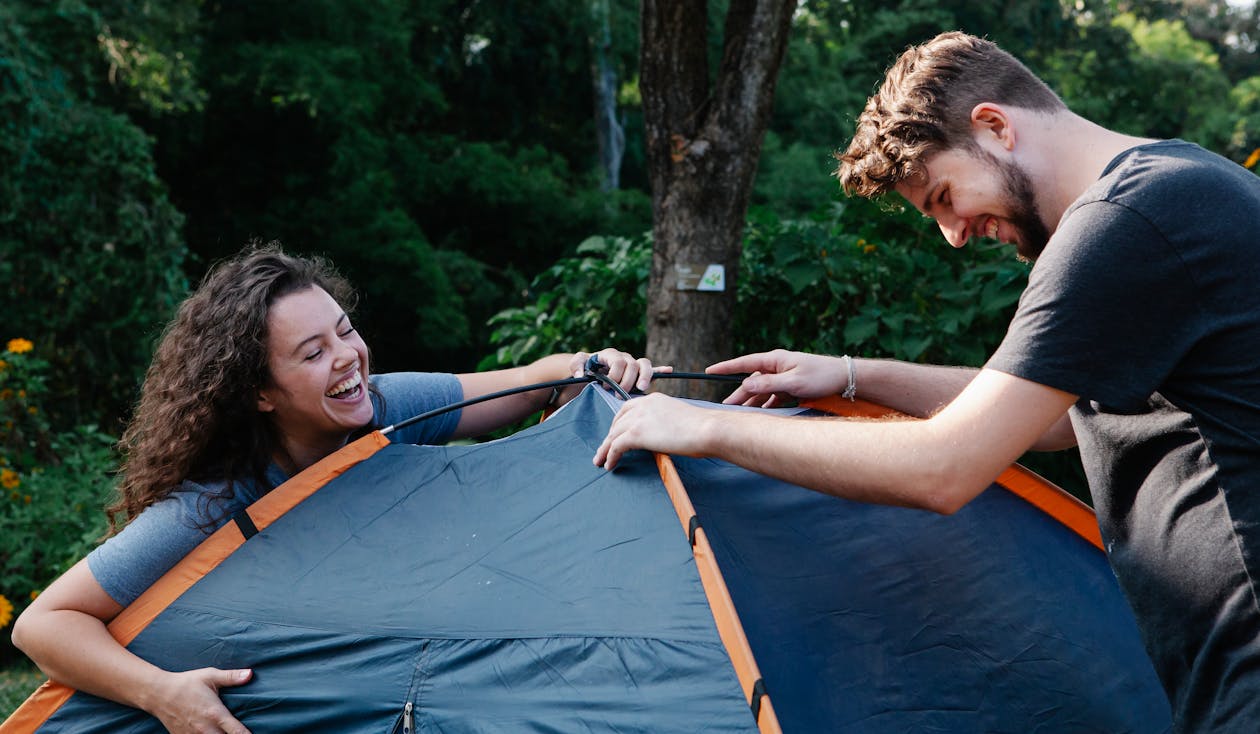 Cheerful young couple in casual clothes laughing while setting up camping tent during romantic picnic in nature