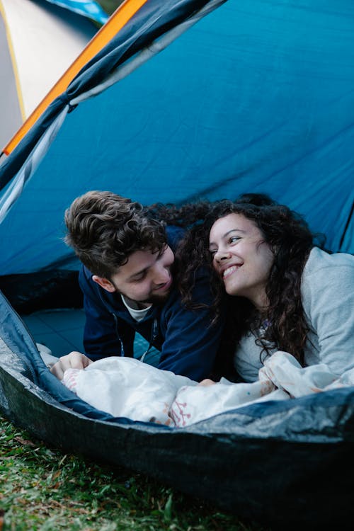 Happy loving young couple in warm clothes smiling and looking at each other while recreating in tent during camping
