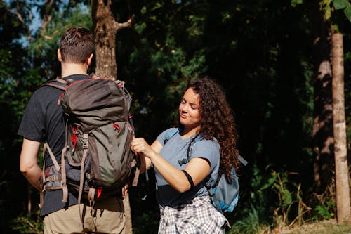 Free Young female hiker in casual clothes fastening backpack of unrecognizable boyfriend during trekking in lush green forest on sunny day Stock Photo