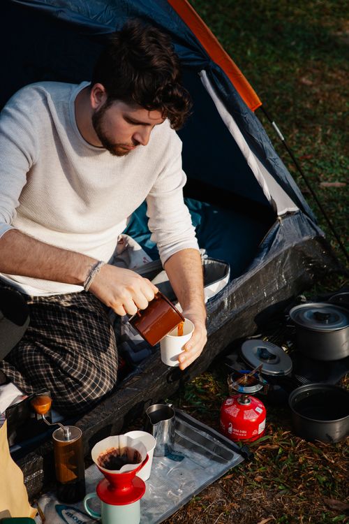 Free From above of peaceful young male camper in casual clothes pouring fresh brewed coffee into cup while relaxing in tent on sunny day Stock Photo