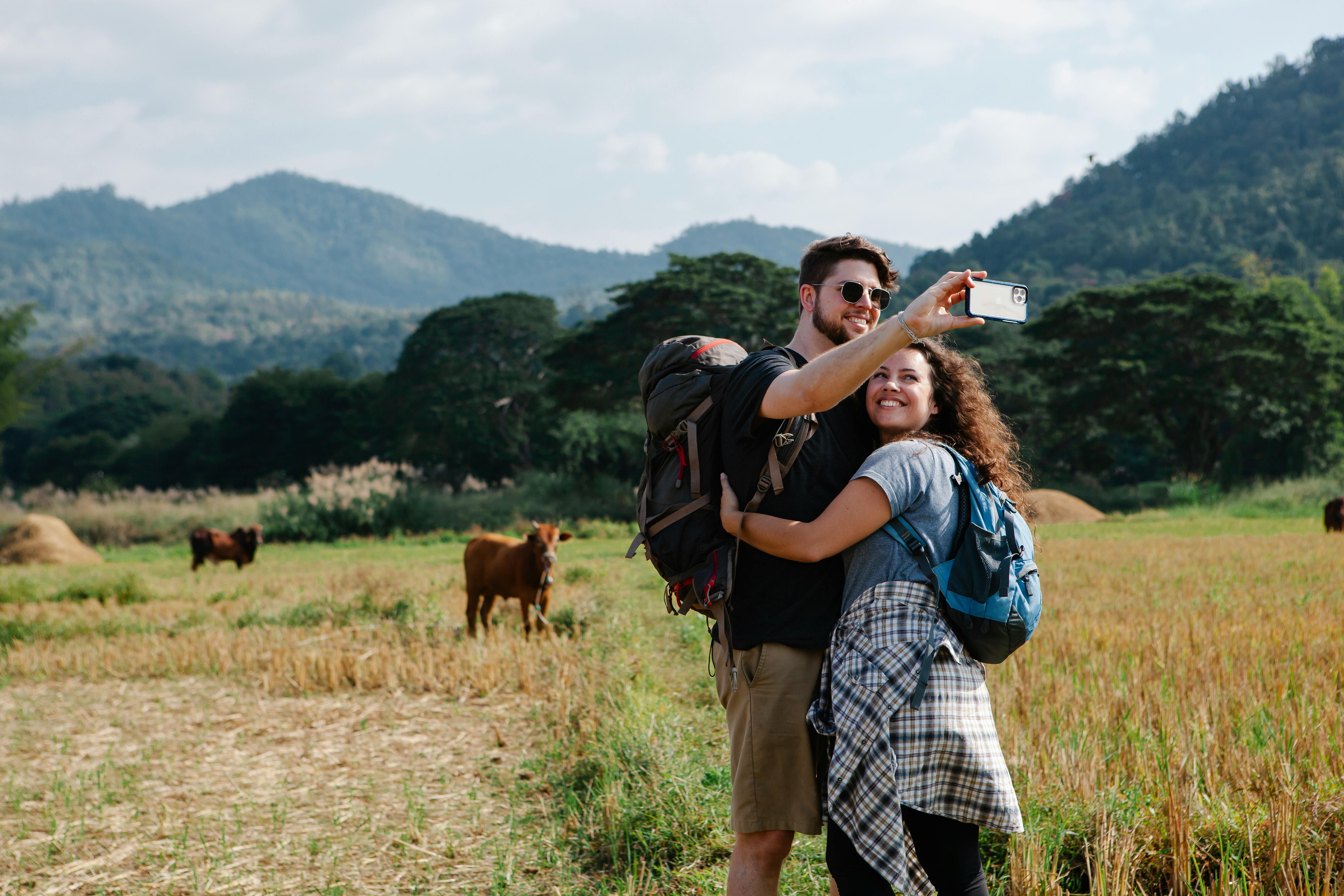 happy couple of travelers hugging and taking selfie in field against hills