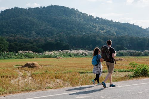 Back view of anonymous couple of travelers in casual clothes with backpacks walking along asphalt roadway against green field and hills placed in countryside in sunny day