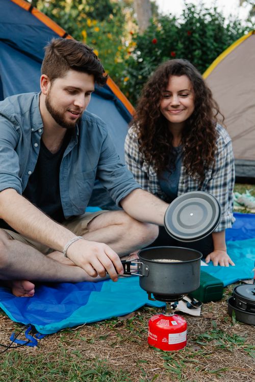 Free Positive couple of hikers in casual clothes sitting on ground near tents and preparing food in pot while traveling in nature Stock Photo