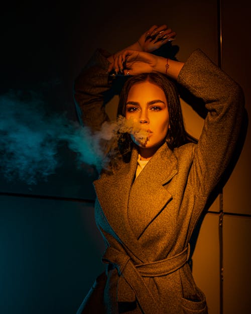 Stylish woman exhaling smoke in light