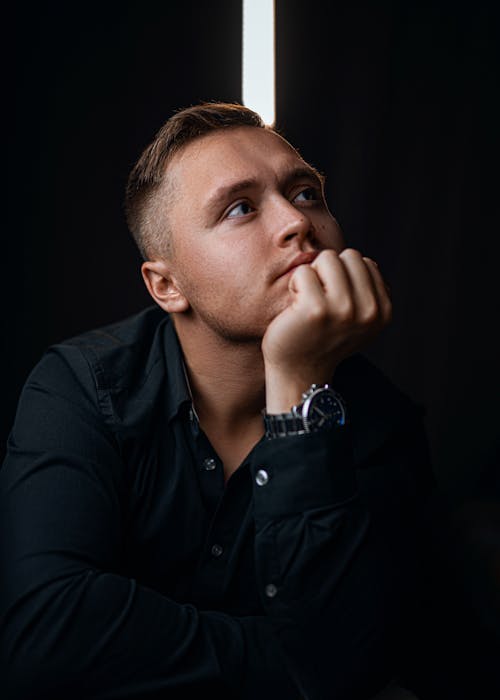 Pensive young male with wristwatch in formal wear touching chin while sitting in dark room and looking away