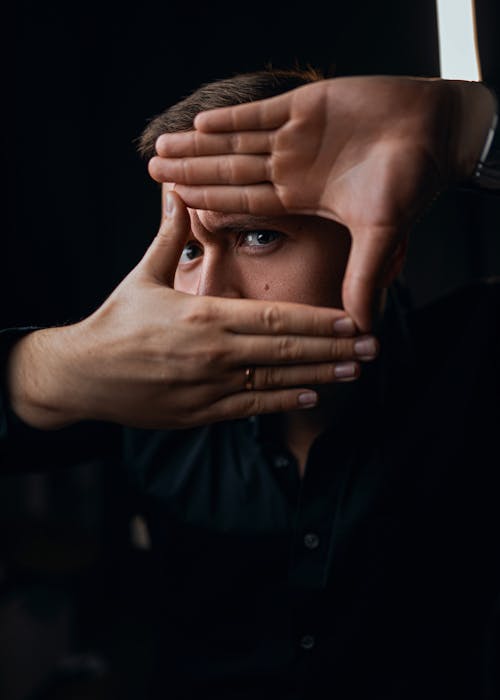 Frowning male with ring covering face with hand while standing in dark room and looking at camera