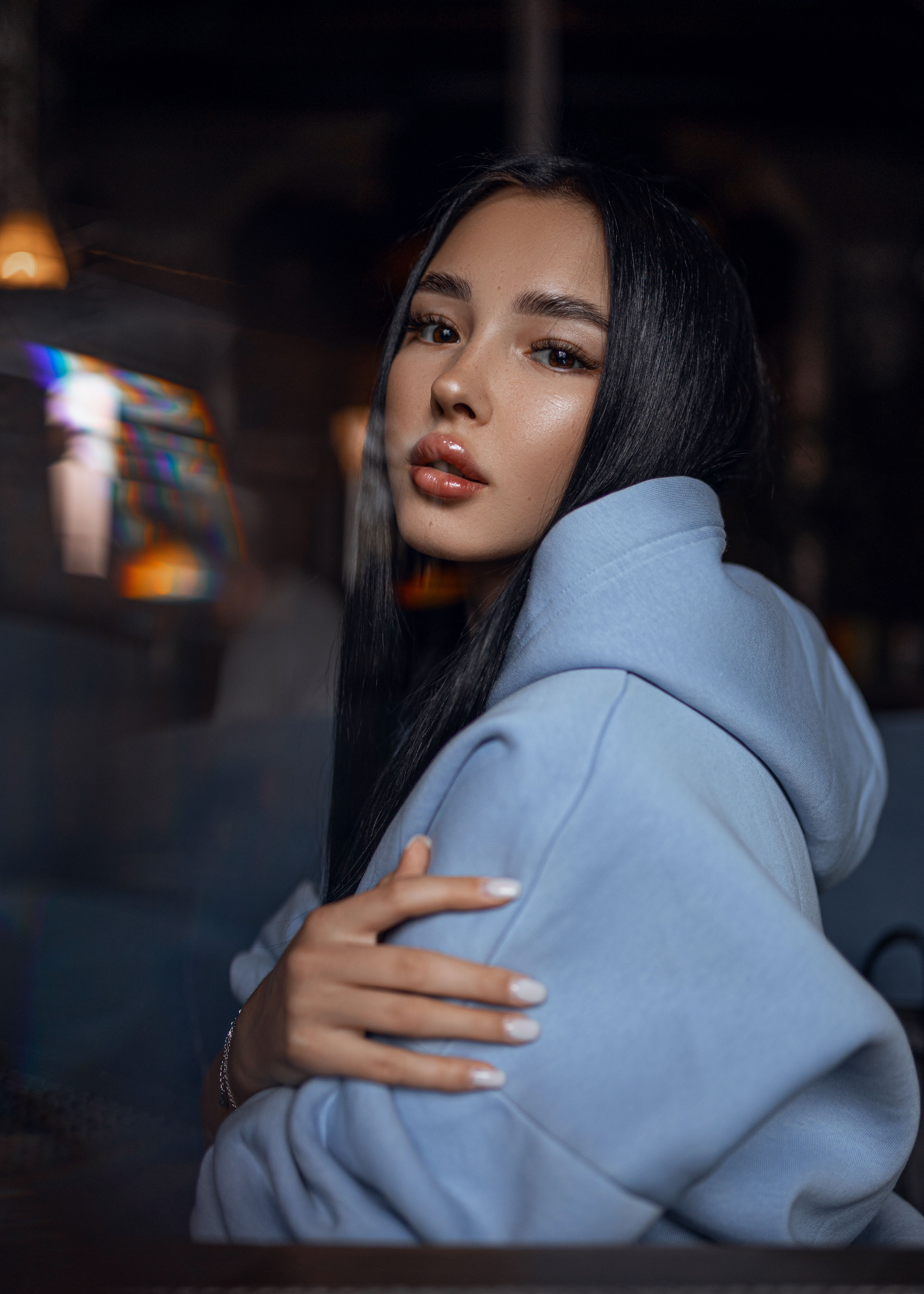 Side view of young female with long dark hair and makeup in hoodie sitting at table and looking at camera