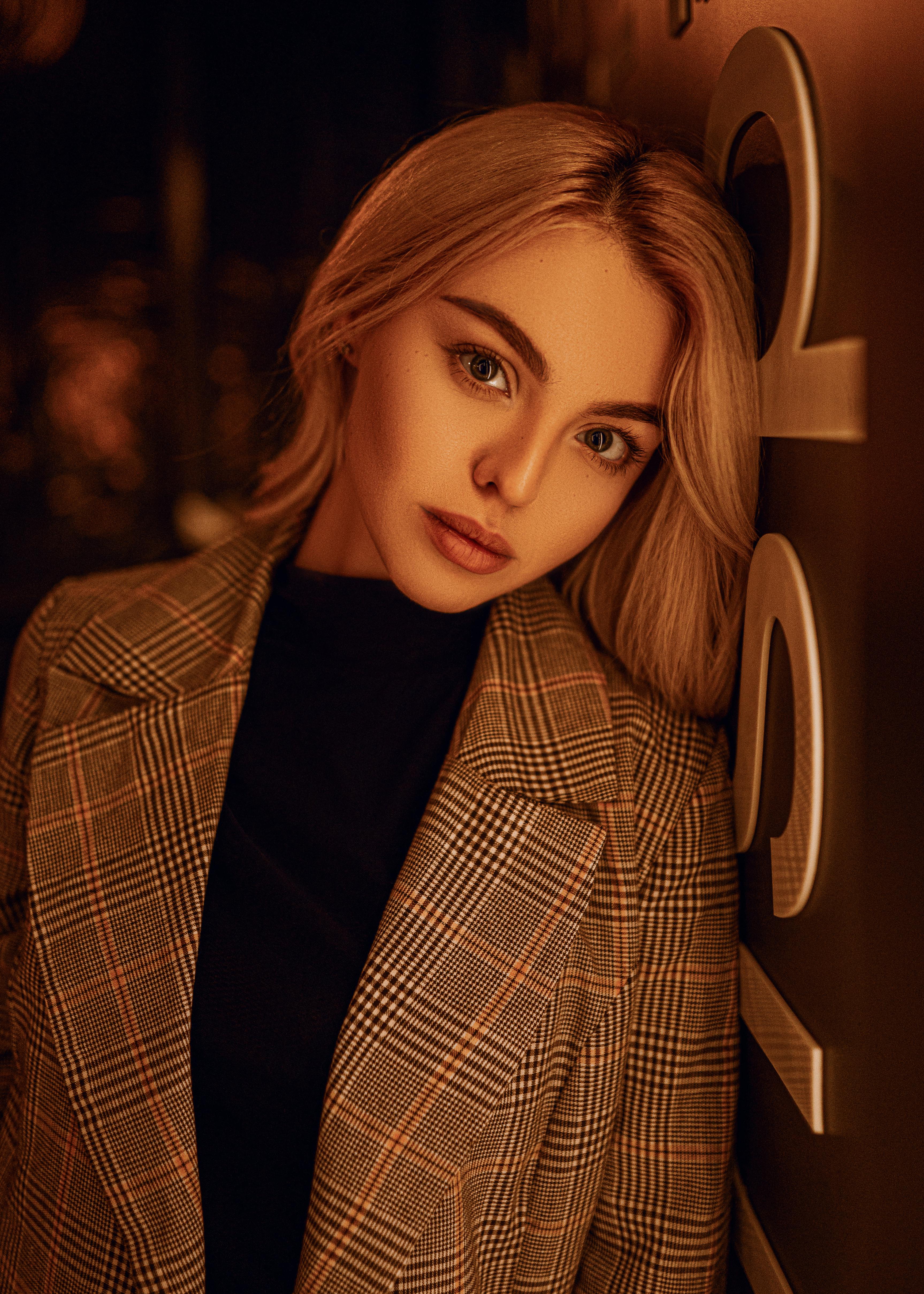 Young charming female with blond hair and light makeup in stylish coat standing near wall and looking at camera