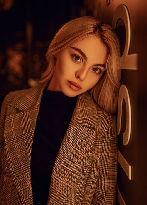 Free Young charming female with blond hair and light makeup in stylish coat standing near wall and looking at camera Stock Photo