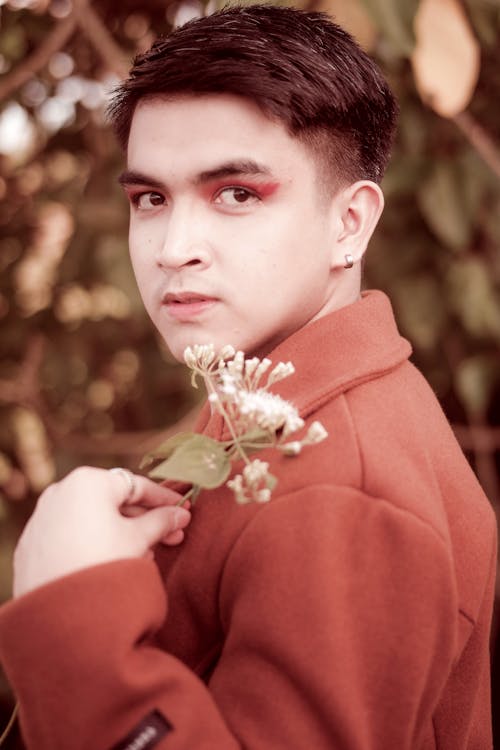 Side view of dark haired young male model with eyeshadow holding bunch of flowers and looking at camera