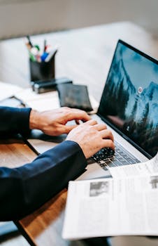 Professional working on a laptop with papers and phone on a wooden table indoors.