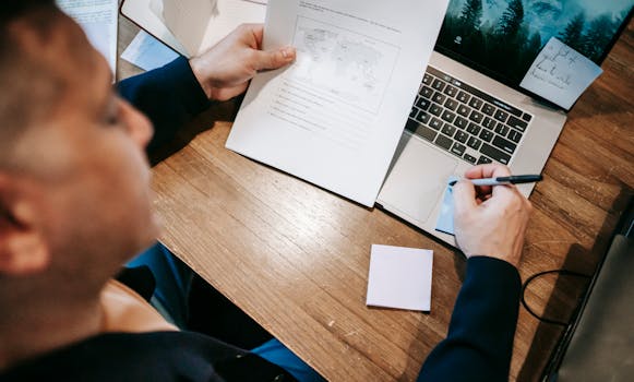 Man engaged in remote work, using laptop and analyzing documents at a wooden table.