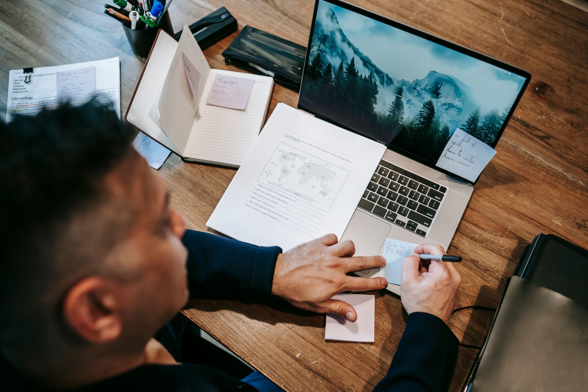 Man working remotely on laptop at a wooden desk with papers, notes, and a notebook.
