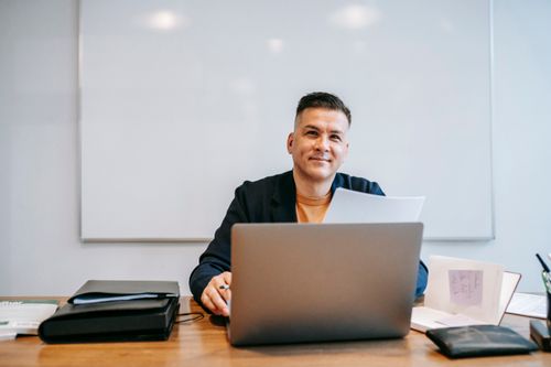 Free Confident man working remotely with a laptop at home office, smiling at his workspace. Stock Photo