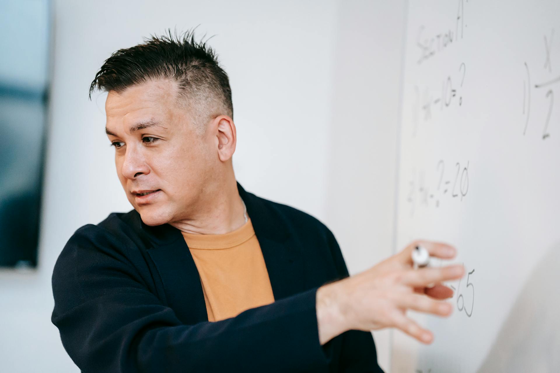 A male teacher explains mathematical concepts on a whiteboard in a classroom setting.