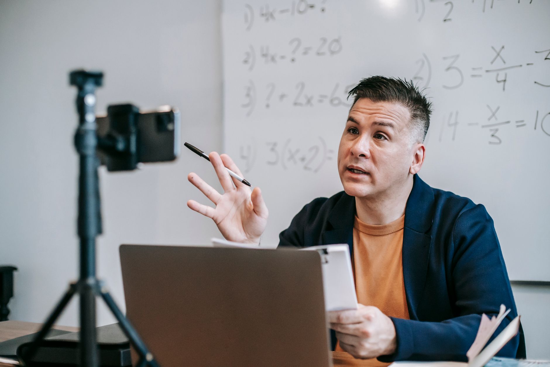 Ein Mann, der vor einem Laptop und einer Kamera auf einem Stativ spricht, im Hintergrund ein Whiteboard.