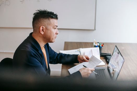 Side view of serious adult guy in casual clothes sitting on chair at table and having video call on netbook with colleague with papers and pen in light room