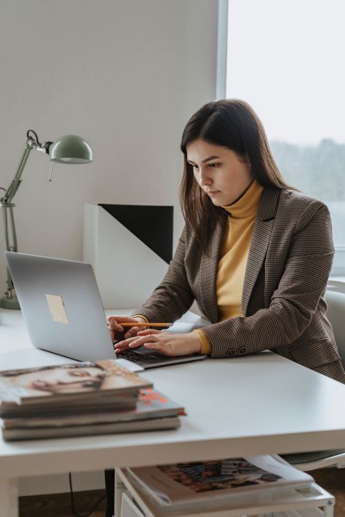 Free Businesswoman typing on laptop in modern office workspace with a desk lamp and magazines. Stock Photo