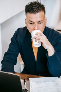 Businessman drinking coffee while working on laptop at home office desk.