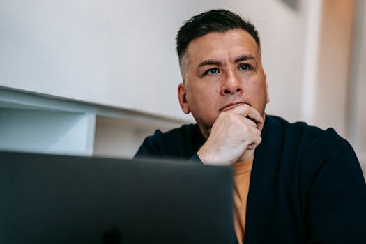 Focused man in home office pondering work on a laptop, expressing deep concentration and thought.