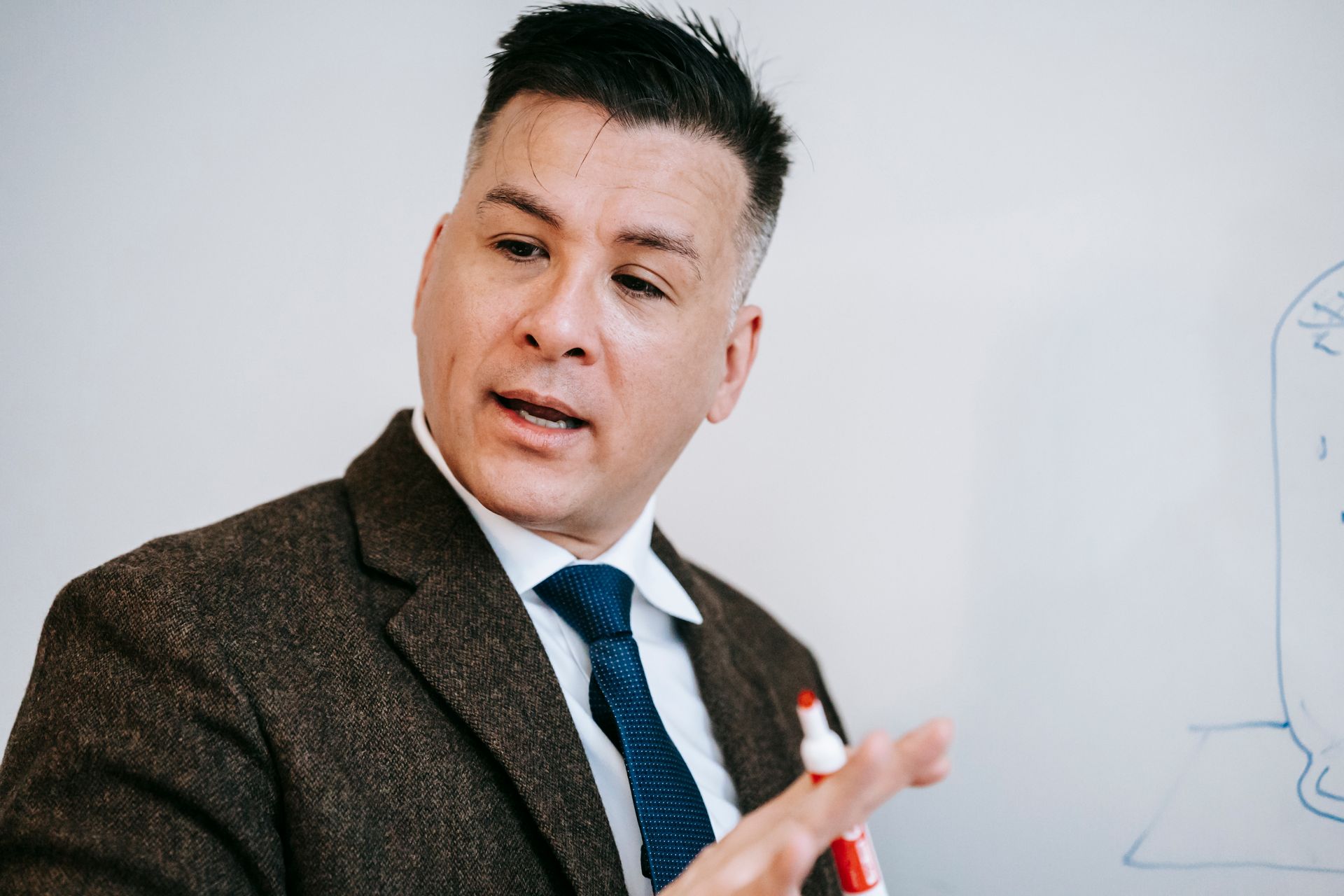 A professional male educator in formal attire using a marker to explain a concept on a whiteboard.