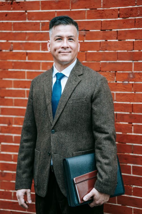 Free Businessman in formal attire against a red brick wall, holding a briefcase with a confident smile. Stock Photo