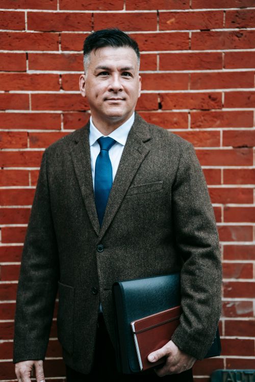 Free Confident businessman in formal attire standing against a brick wall holding notebooks. Stock Photo