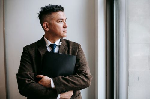 Free A thoughtful man in formal attire holding a notebook, gazing out of a window. Stock Photo