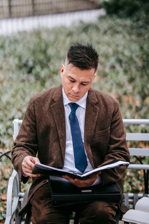 Free Concentrated adult male in elegant clothes sitting on bench and reading papers in folder near notebook in park near grassy lawn in city in daytime Stock Photo
