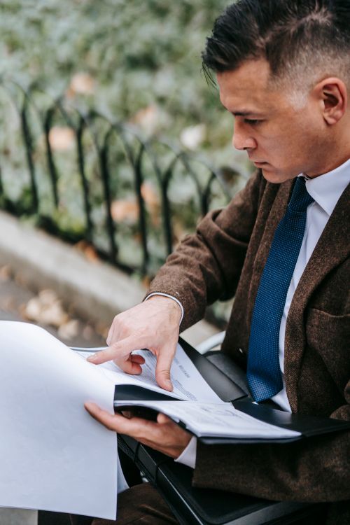 Free A professional man in formal attire reads documents while sitting outdoors, demonstrating focused business engagement. Stock Photo