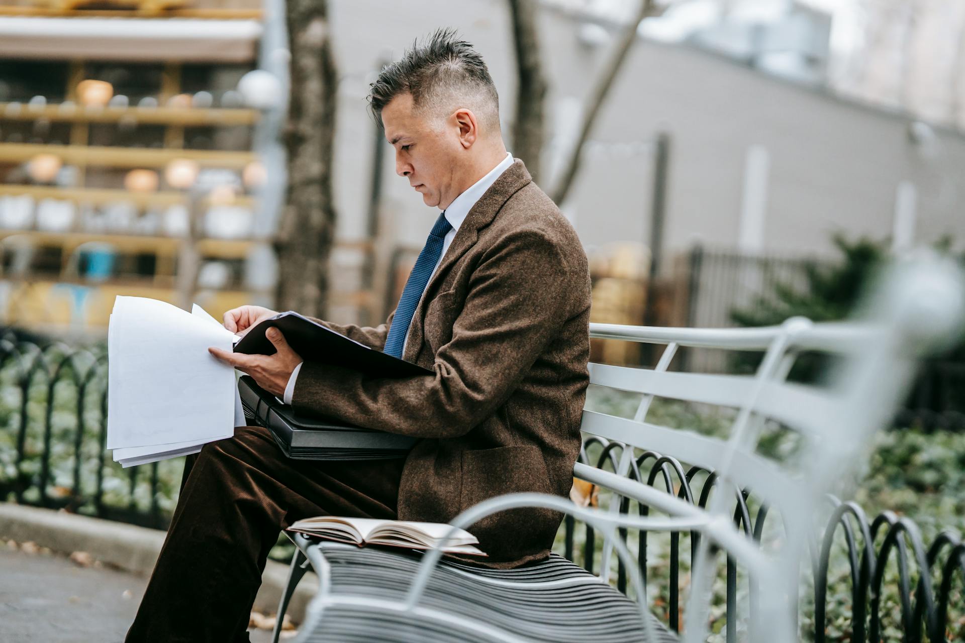 A professional man sitting on a park bench reviewing documents. Business attire, focused and engaged.