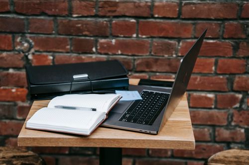 Free Open laptop and notebook on a wooden table by a brick wall, perfect for remote work inspiration. Stock Photo