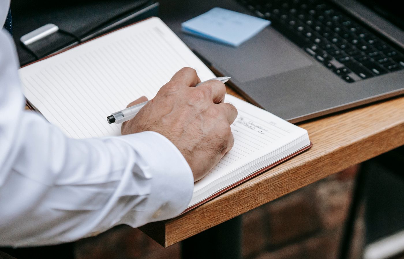 Close-up of an adult writing in a notebook beside a laptop. Professional setting.