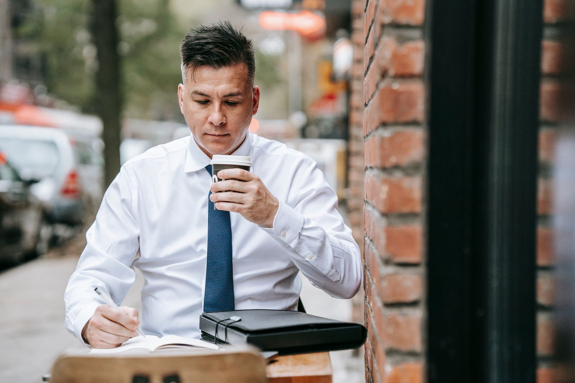 Professional man in corporate attire writing and sipping coffee outdoors on a bustling city sidewalk.