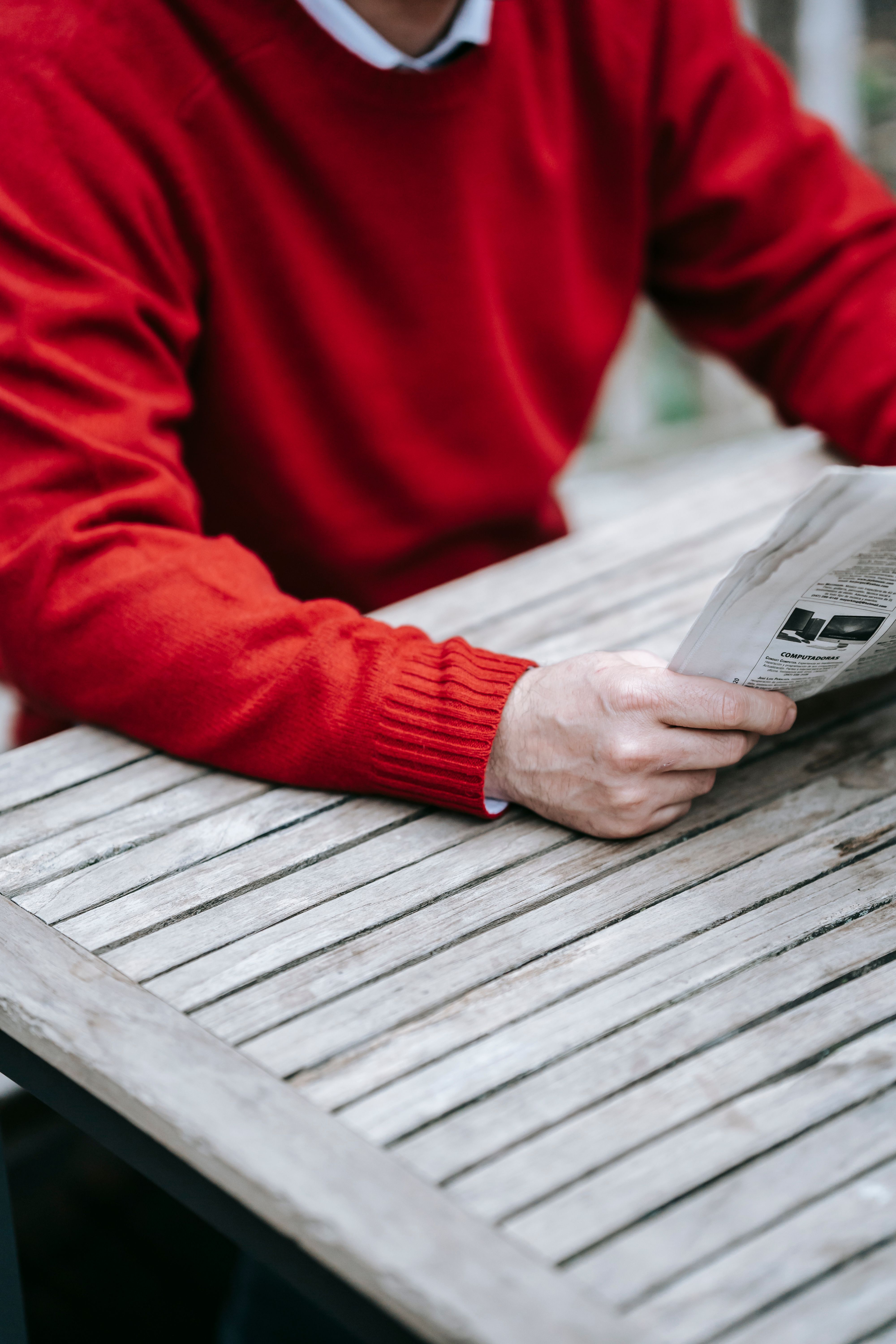 Person in red sweater reads newspaper at a wooden table, relaxed ambiance.
