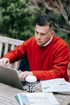 A man in a red sweater working remotely on a laptop at a wooden table outdoors.