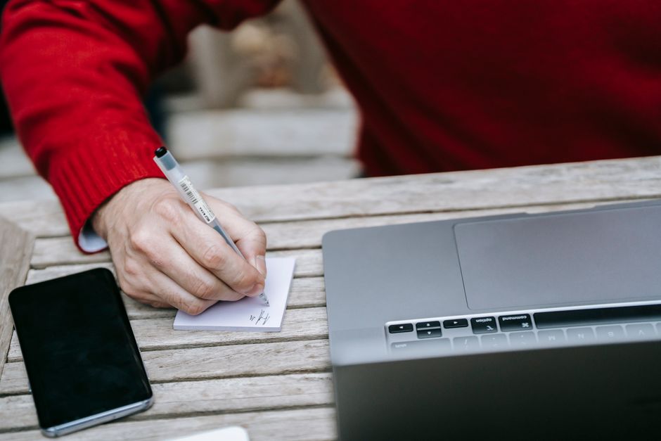 Adult writing notes beside a laptop and smartphone on a wooden table outdoors.