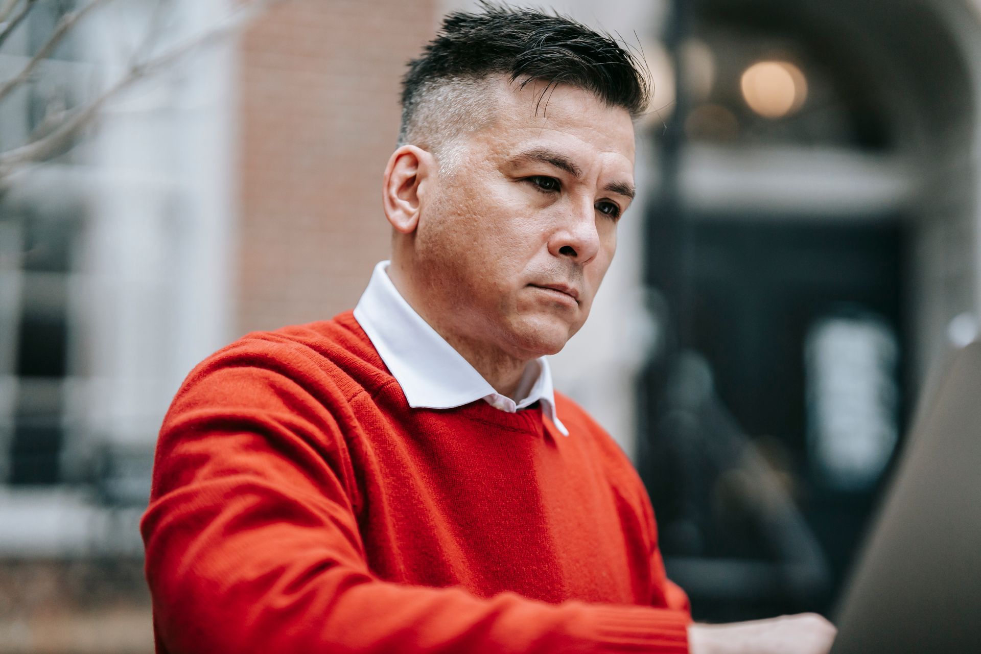 Serious man in red sweater focused on work using laptop outside an office building.