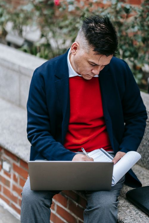 Free A stylish man working on a laptop and taking notes outside, embodying remote work lifestyle. Stock Photo