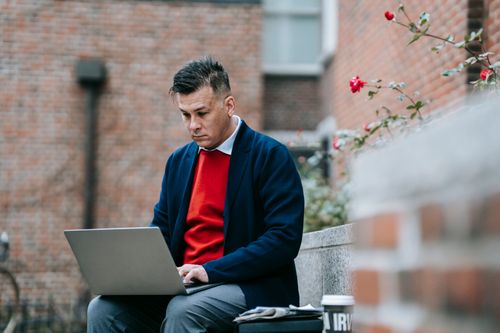 Free Businessman working remotely on a laptop in an outdoor brick-setting with flowers. Stock Photo